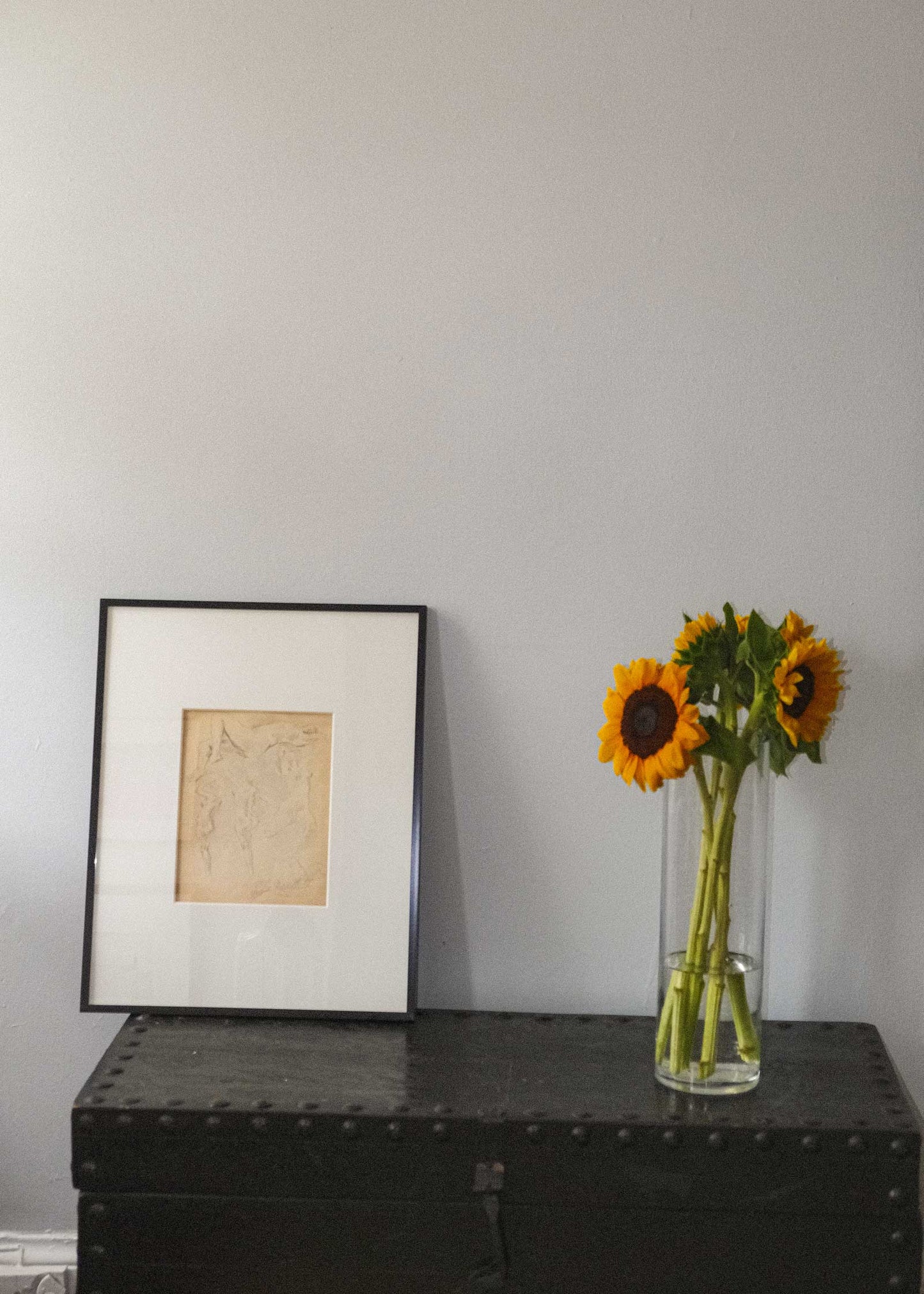 Framed artwork and a vase with sunflowers on a dark surface against a light gray wall.