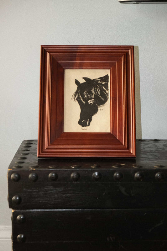 Framed artwork of a horse's head on a wooden surface with a white wall background