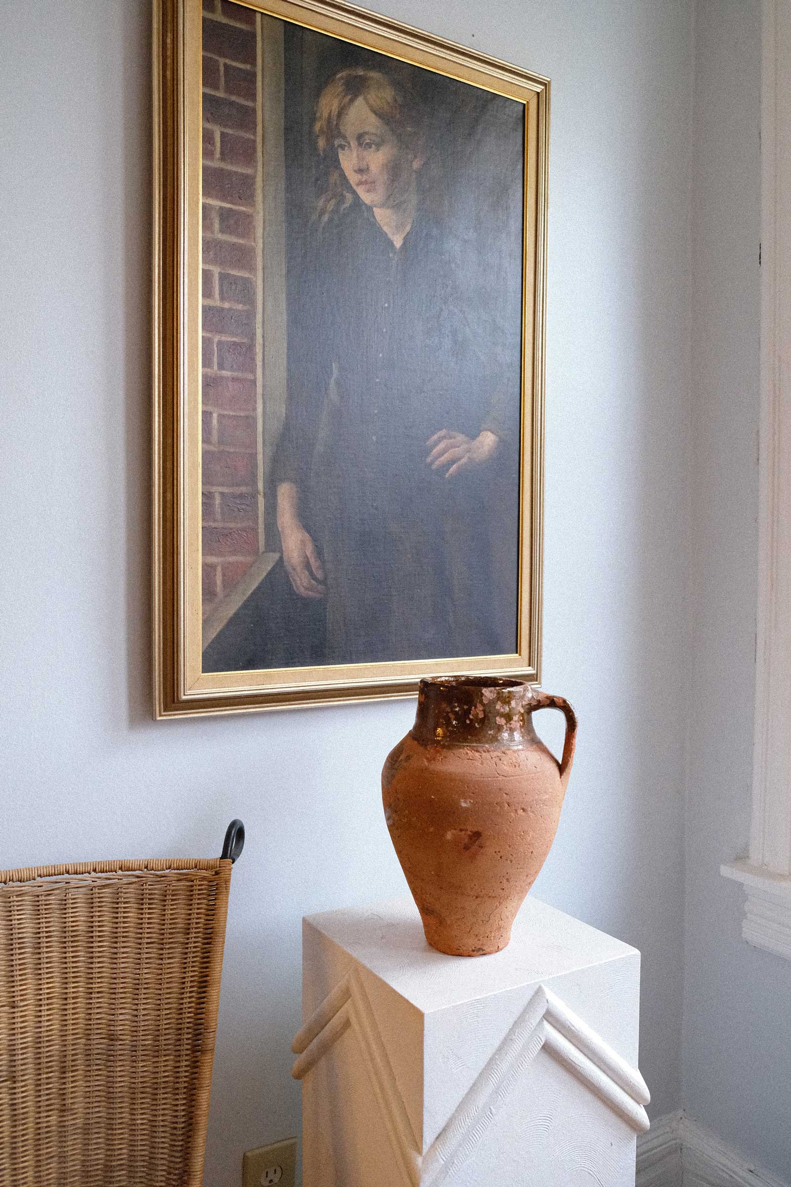 Framed portrait on a wall with a terracotta vase on a pedestal below.