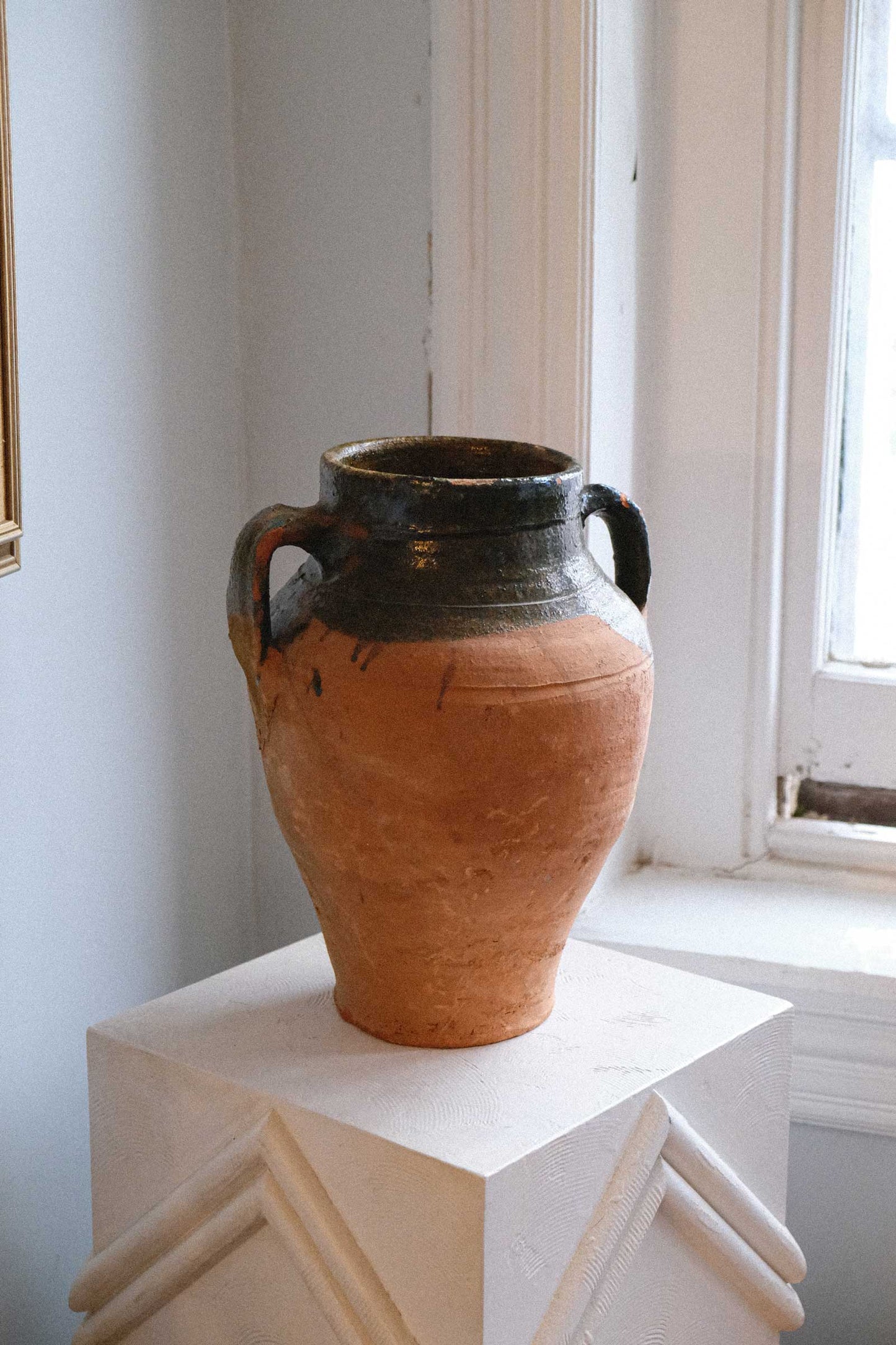 Terracotta vase on a white pedestal against a light-colored wall with a window.