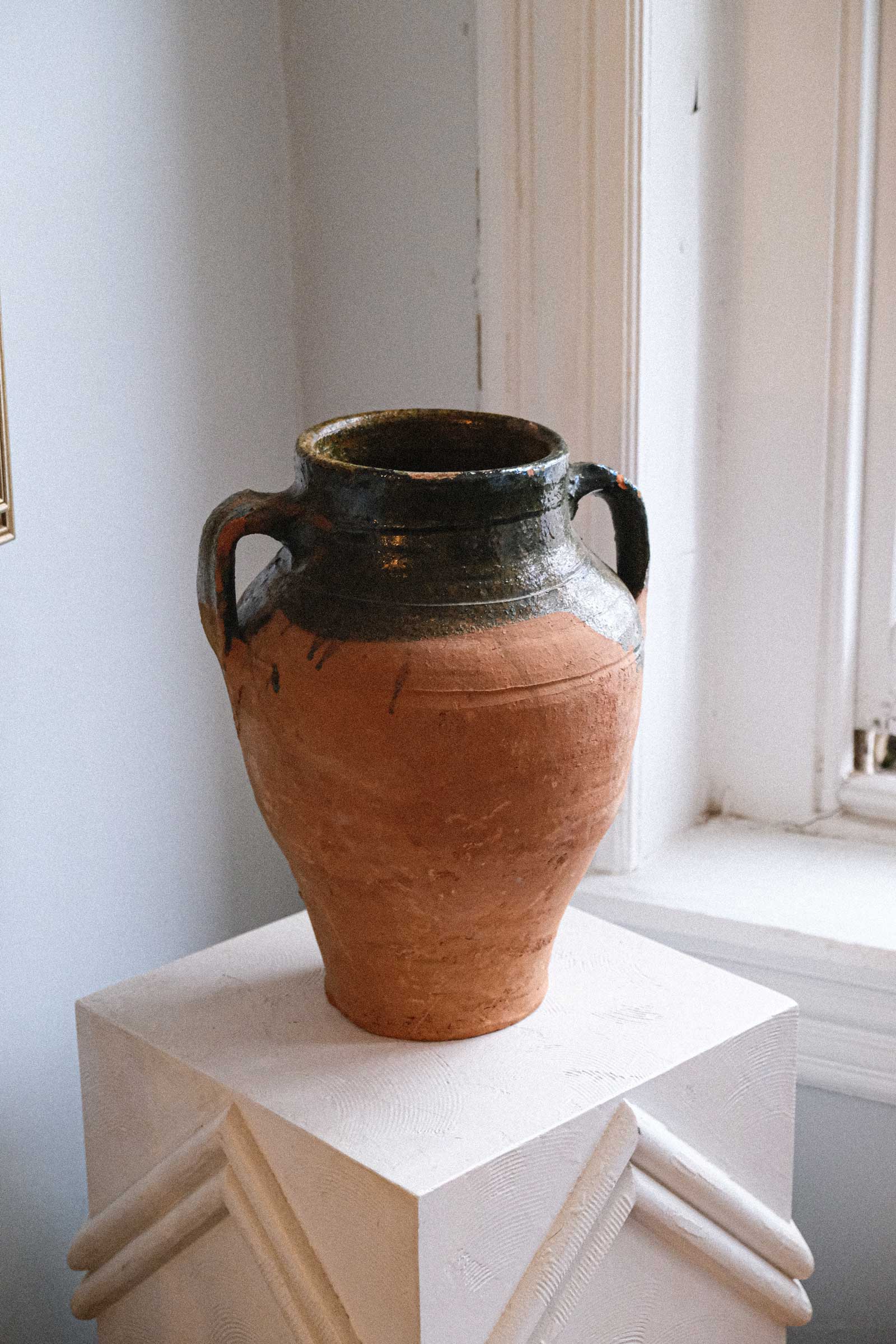 Terracotta vase with black glaze on a white pedestal against a light-colored wall.